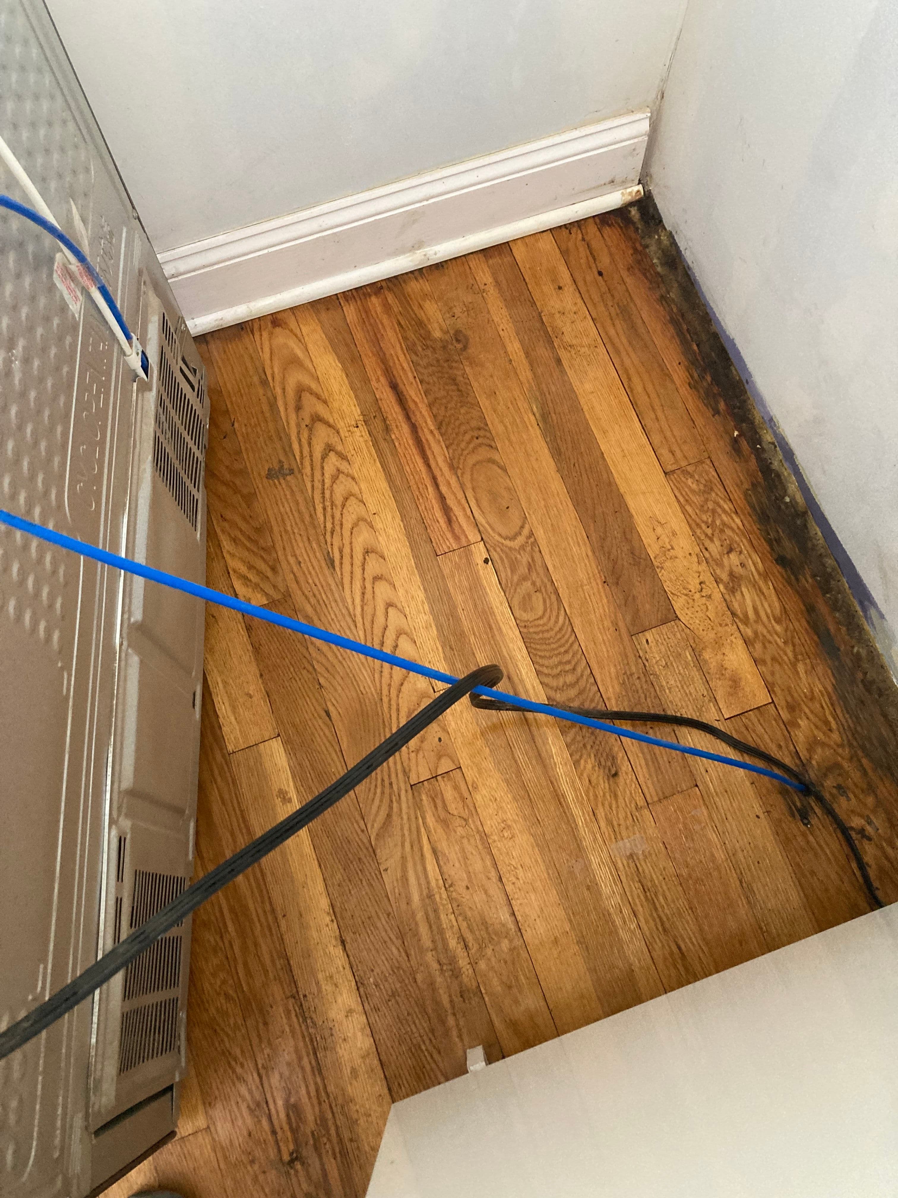 Worn wooden floor with visible water damage and electrical cables near an appliance.