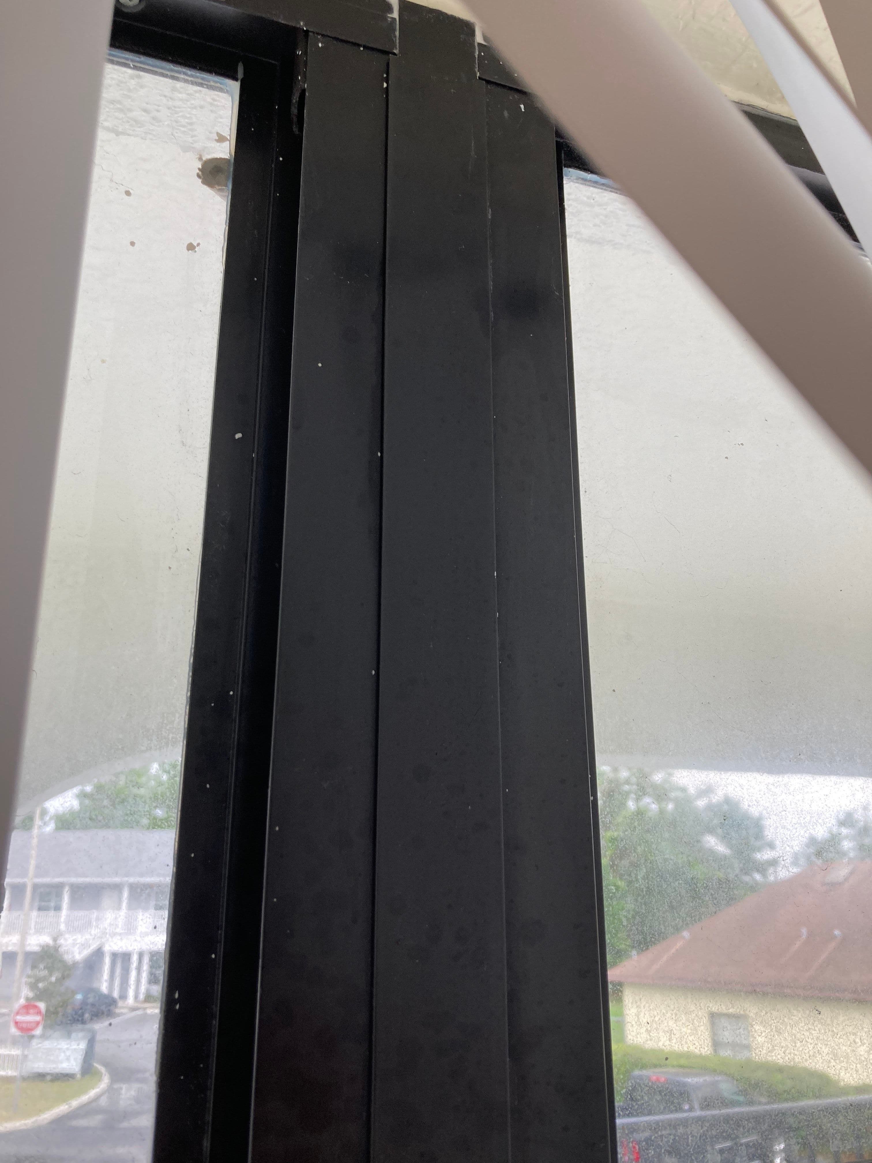 Black window frame with vertical slats, sunlight filtering through dusty blinds.